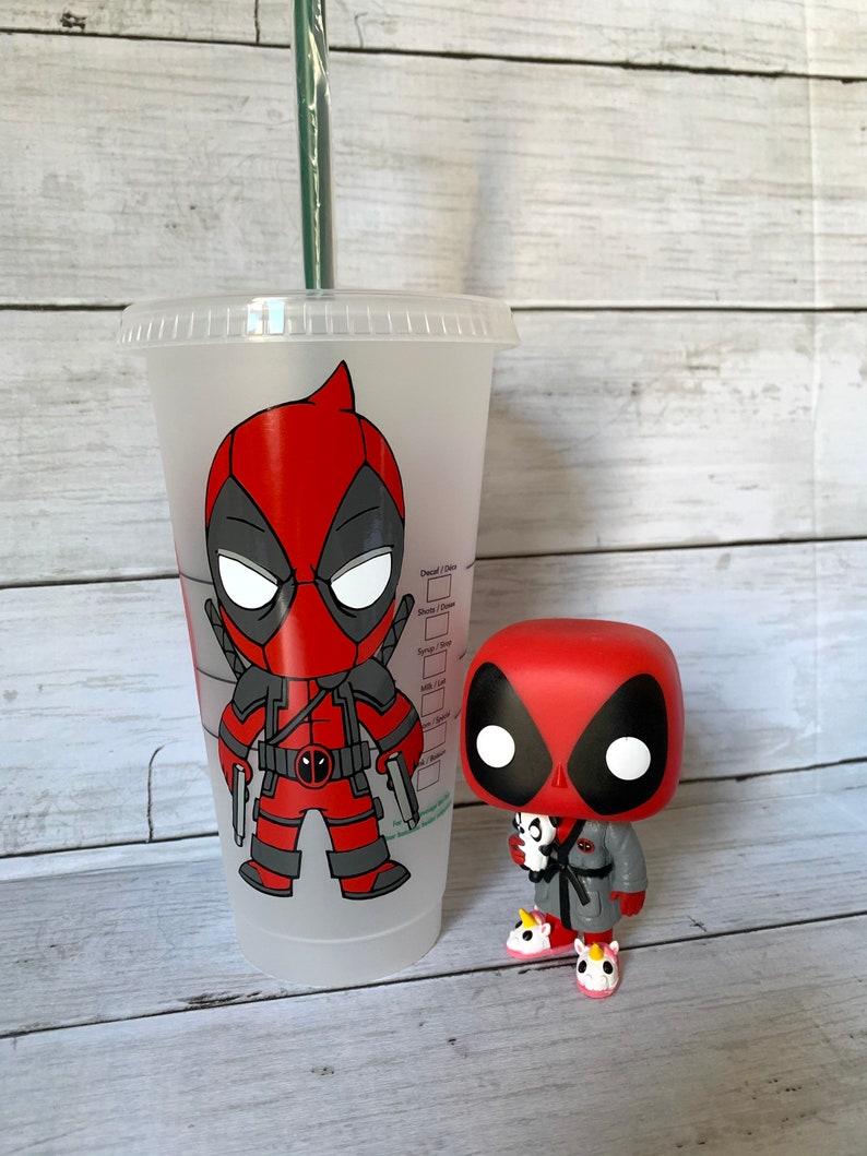 以下が含まれることがあります： 透明なプラスチック製の再利用可能なカップで、赤と黒のカートゥーンのデッドプールキャラクターのデザインが施されています。カップの隣には、より小さな赤と黒のデッドプールフィギュアが立っています。