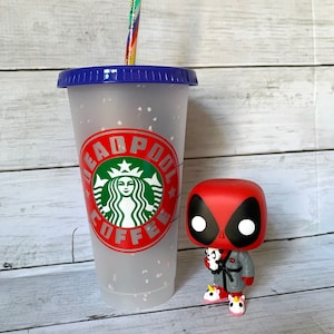 以下が含まれることがあります： 青い蓋と虹色のストローが付いた、白いフロスト加工の再利用可能なカップ。カップには、緑色のスターバックスのロゴと、「DEADPOOL COFFEE」という赤い文字が印刷されています。カップの隣には、赤と黒のデッドプール・ファンコポップフィギュアが立っています。