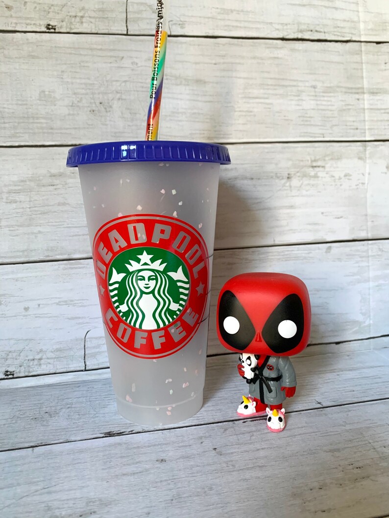 以下が含まれることがあります： 青い蓋と虹色のストローが付いた、白いフロスト加工の再利用可能なカップ。カップには、緑色のスターバックスのロゴと、「DEADPOOL COFFEE」という赤い文字が印刷されています。カップの隣には、赤と黒のデッドプール・ファンコポップフィギュアが立っています。