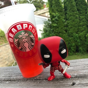 以下が含まれることがあります： 赤い黒のデッドプールファンコポップフィギュアが、銀のナイフを2本持っています。フィギュアは、デッドプールのロゴが入った赤いスターバックスカップの隣の木製表面に立っています。