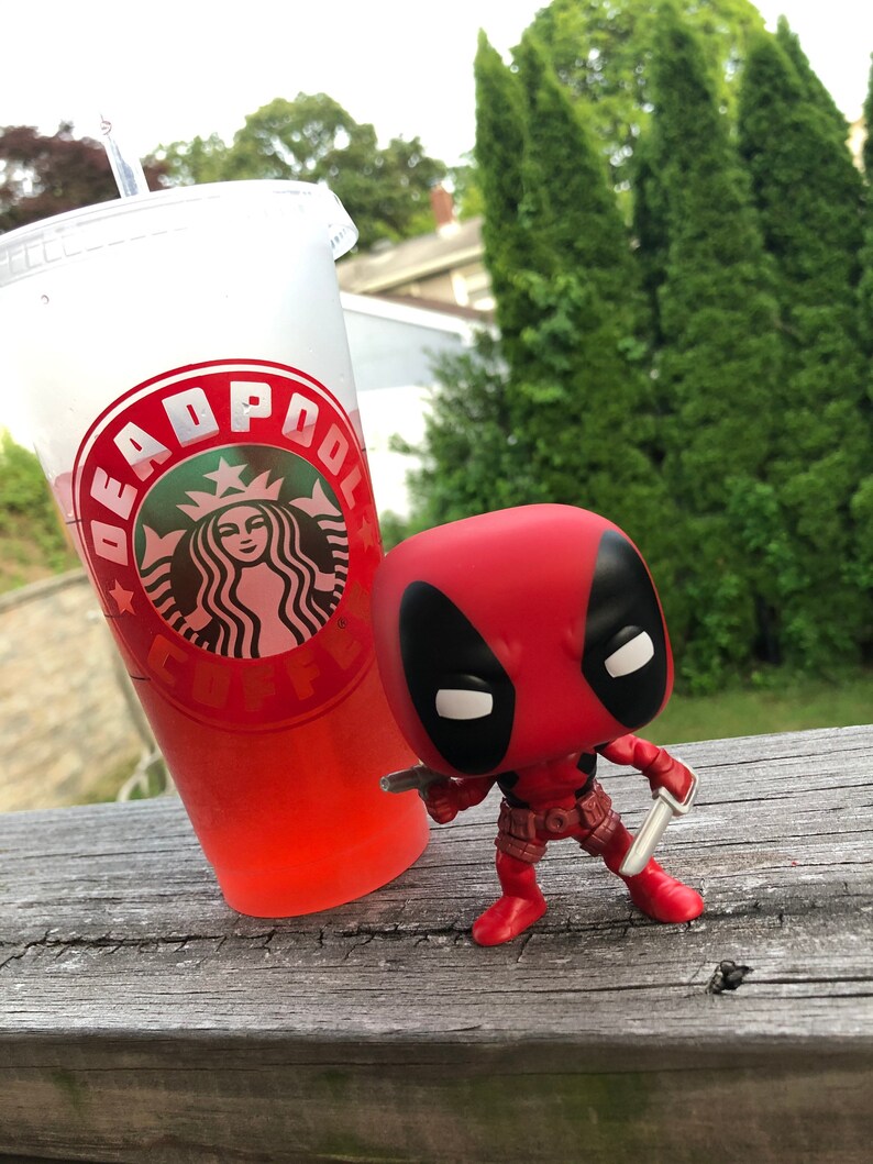 以下が含まれることがあります： 赤い黒のデッドプールファンコポップフィギュアが、銀のナイフを2本持っています。フィギュアは、デッドプールのロゴが入った赤いスターバックスカップの隣の木製表面に立っています。