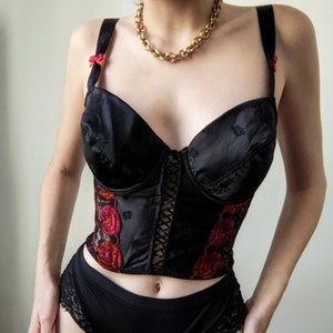 May include: Black satin corset with red and white floral embroidery and a black lace trim. The corset has a busk closure in the front and adjustable straps.