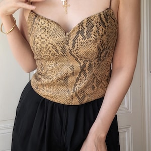 May include: A sleeveless corset-style top featuring a brown and black snakeskin pattern with thin spaghetti straps and a sweetheart neckline. The garment is paired with solid black trousers, a gold chain necklace with a cross pendant, and a gold ring.