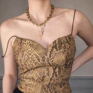 Könnte beinhalten: Ein braunes und schwarzes Oberteil mit Schlangenhautmuster, Sweetheart-Ausschnitt und dünnen Trägern. Das Oberteil wird mit einer goldenen Kettenkette mit einem Kreuz- und einem Ankh-Anhänger getragen.