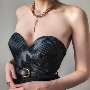 May include: A black strapless top with a pleated bodice and a gold chain necklace with a cross pendant. The top is paired with black pants and a gold belt.