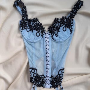 May include: Light blue mesh corset with black floral lace detailing. Features adjustable straps, a hook-and-eye closure, and garter straps with satin bows. The corset is designed with a sweetheart neckline and a delicate, feminine aesthetic.