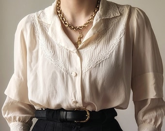 May include: A white, long-sleeved blouse with delicate lace detailing around the collar and cuffs. The blouse is worn with a black high-waisted skirt and a gold chain necklace.