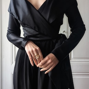 May include: Black wrap-style jumpsuit with long sleeves. The outfit features a V-neckline and a matching belt. A gold-colored necklace and ring are also visible. The jumpsuit is made of a shiny, silky material.