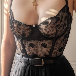 K&ouml;nnte beinhalten: Schwarzes Korsett aus floraler Spitze mit herzf&ouml;rmigem Ausschnitt, kombiniert mit einem schwarzen G&uuml;rtel und einer hoch taillierten schwarzen Hose. Eine goldene Halskette mit Kreuzanh&auml;nger erg&auml;nzt das Outfit. Das Korsett hat zarte Blumenstickereien.