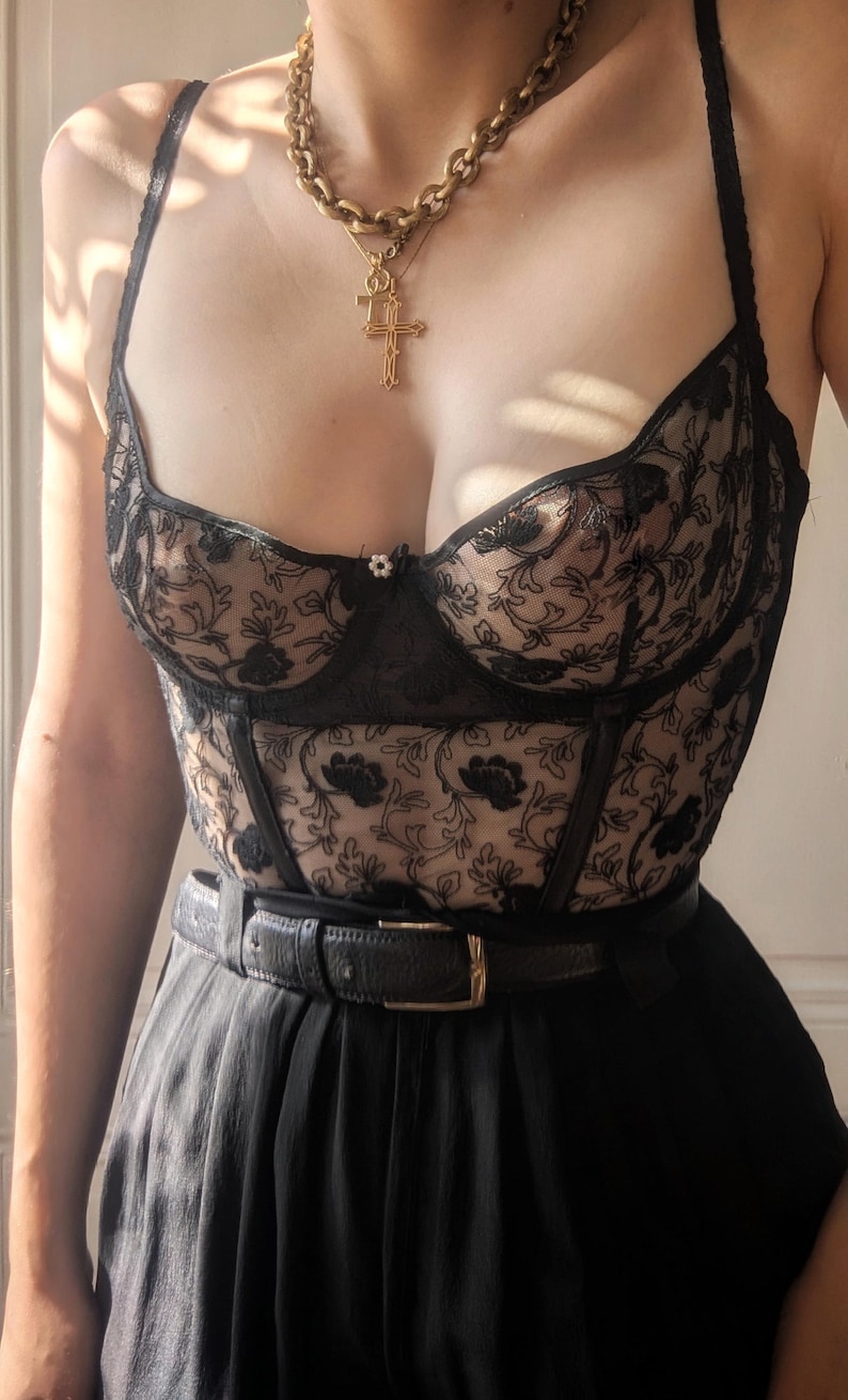 K&ouml;nnte beinhalten: Schwarzes Korsett aus floraler Spitze mit herzf&ouml;rmigem Ausschnitt, kombiniert mit einem schwarzen G&uuml;rtel und einer hoch taillierten schwarzen Hose. Eine goldene Halskette mit Kreuzanh&auml;nger erg&auml;nzt das Outfit. Das Korsett hat zarte Blumenstickereien.