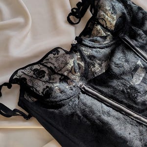 K&ouml;nnte beinhalten: Schwarzes Spitzenkorsett mit floralen Details. Das Korsett hat ein strukturiertes Oberteil mit St&auml;bchen und zarten Spitzentrimm. Das Kleidungsst&uuml;ck ist vor einem Hintergrund aus elfenbeinfarbenem Stoff platziert, wodurch ein Kontrast aus Texturen und Farben entsteht.