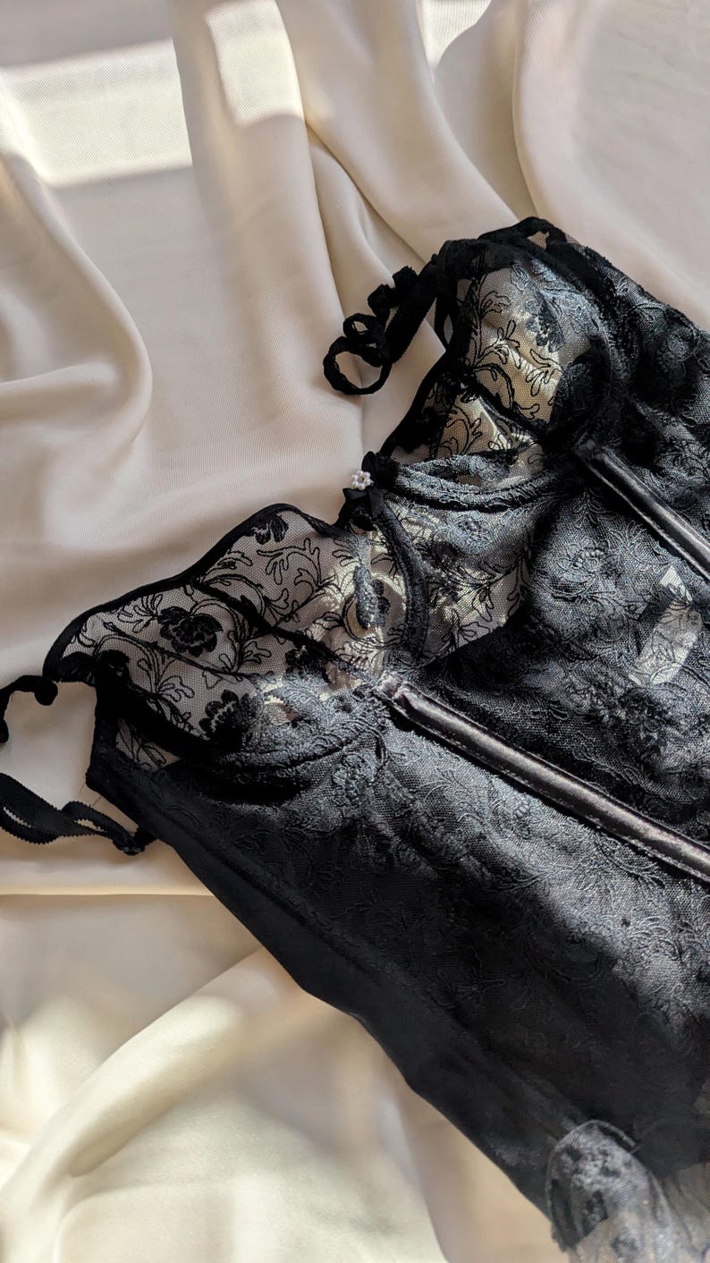 K&ouml;nnte beinhalten: Schwarzes Spitzenkorsett mit floralen Details. Das Korsett hat ein strukturiertes Oberteil mit St&auml;bchen und zarten Spitzentrimm. Das Kleidungsst&uuml;ck ist vor einem Hintergrund aus elfenbeinfarbenem Stoff platziert, wodurch ein Kontrast aus Texturen und Farben entsteht.