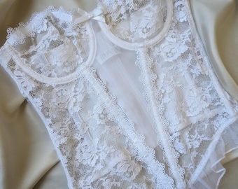May include: White lace corset with a delicate floral pattern. The corset features a sweetheart neckline, a small bow, and a ruffled hem. The garment is made of a soft, sheer fabric and is displayed on a satin surface.