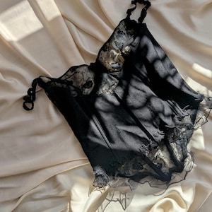 K&ouml;nnte beinhalten: Schwarzes Dessous-Top mit zarten Spitzendetails. Das Top hat verstellbare Tr&auml;ger und einen ger&uuml;schten Saum. Der Stoff wirkt weich und drapiert sich &uuml;ber einer cremefarbenen Oberfl&auml;che, wobei Schatten dem Bild Tiefe verleihen.