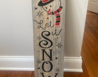 May include: A wooden sign with a snowman design and the text "Let it Snow" painted in black. The snowman is wearing a black top hat with a red band and a red and black scarf.