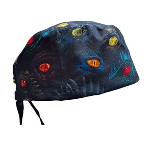 May include: A black scrub cap featuring a repeating pattern of reptilian eyes in various colors including yellow, orange, red, and blue. The fabric displays detailed scales surrounding each eye, with tie-back straps visible at the bottom edge.