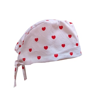 May include: A white scrub cap featuring a repeating pattern of small red hearts. The head covering has a rounded crown and includes two fabric ties at the back for securing the fit, providing a functional and simple design for medical or clinical use.