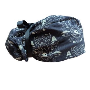May include: A dark blue scrub cap with a repeating pattern of white mushrooms, eyes, and other whimsical elements. The cap is tied with a black ribbon. The fabric has a soft, matte finish.