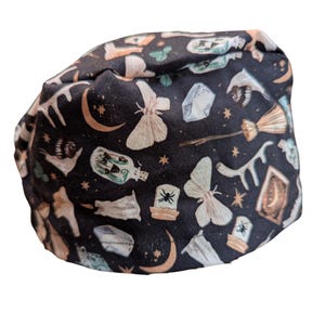 May include: A black fabric item with a repeating pattern of celestial and mystical elements. The design includes moons, stars, moths, crystals, potion bottles, and a broom. The color palette features black, cream, and gold.