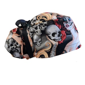 May include: A patterned scrub cap featuring a design with skulls, roses, and a woman with sugar skull makeup. The cap has a black ribbon tie and is made of a soft, flexible fabric. The color palette includes black, gray, red, and tan.