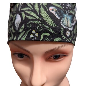 May include: Black fabric head covering with a repeating pattern of green ferns, mushrooms, a moth, an owl, and a skull. The head covering is worn by a mannequin.
