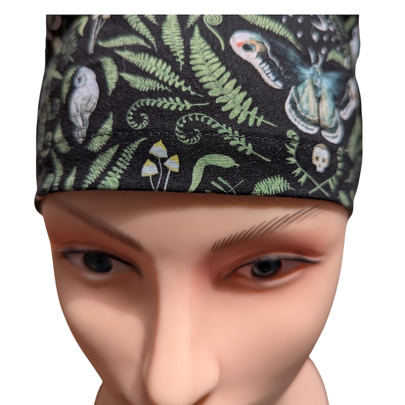 May include: Black fabric head covering with a repeating pattern of green ferns, mushrooms, a moth, an owl, and a skull. The head covering is worn by a mannequin.