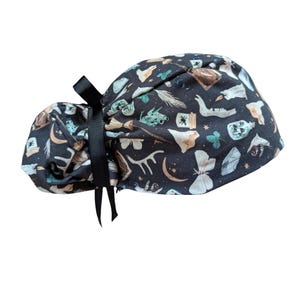 May include: A dark navy blue scrub cap with a black bow. The cap is patterned with various mystical and magical elements in shades of beige, teal, and white. The design includes moons, moths, and potion bottles.