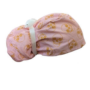 May include: Pink surgical scrub hat with a gold skull pattern. The hat has a white tie.