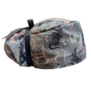 May include: A rolled-up, gray fabric item with a black strap. The fabric is patterned with black cats, witch hats, moons, and other whimsical illustrations. The overall design has a Halloween or fantasy theme.