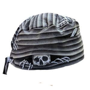 May include: A black and gray skull cap with a repeating pattern of horizontal stripes and skeleton hands. A white skull is featured prominently in the center of the cap. The cap has a tie closure.