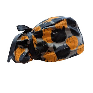 May include: A Halloween-themed scrub cap featuring a pattern of black cats and orange pumpkins on a gray background. The cap has a dark gray ribbon for tying. The fabric is a soft cotton.