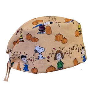 May include: A beige surgical scrub cap featuring a repeating pattern of cartoon characters, pumpkins, and autumn leaves. The characters include Snoopy, Charlie Brown, Lucy, and others. The cap has ties for an adjustable fit.