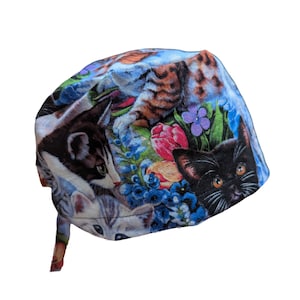 May include: A blue, cat-themed scrub cap featuring a colorful print of various cats, flowers, and foliage. The cap has a tie in the back for an adjustable fit. The design includes a black cat, a tabby cat, and a gray and white cat.
