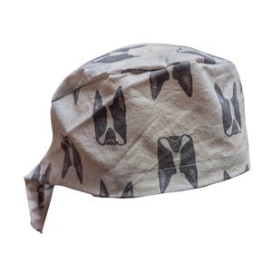 May include: A light beige surgical scrub cap with a repeating pattern of dark gray Boston Terrier dog faces. The cap is tied in the back and is made of a soft fabric.