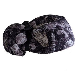 May include: A black and gray patterned scrub hat featuring a full moon, planchette, and Ouija board designs. The hat has a black bow and elastic band for a secure fit. The fabric has a distressed, vintage look.