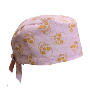 May include: A light pink skull cap with a repeating pattern of gold-colored skulls. The cap is tied at the back with two fabric ties.