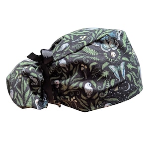 May include: A black scrub cap with a whimsical forest pattern featuring owls, ferns, mushrooms, butterflies, and a crescent moon. The cap has a black bow and is designed to be worn on the head.