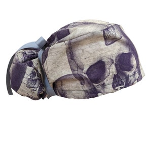 May include: A skull print scrub cap in shades of gray and purple. The cap features a repeating skull design and a light blue ribbon tie. The fabric has a textured appearance, and the overall design is a unique and stylish accessory.