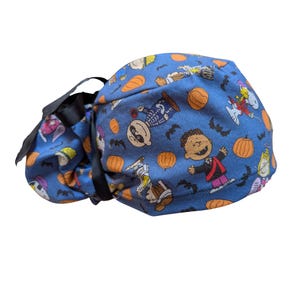 May include: A blue scrub cap featuring a Halloween-themed pattern with cartoon characters, pumpkins, and bats. The cap has a black ribbon tie for adjustment. The fabric is a vibrant blue, and the design is repeated across the surface.
