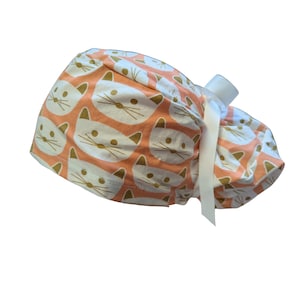 May include: A peach-colored scrub cap with a repeating pattern of white cat faces with gold accents. The cap is tied with a white ribbon. The cap is designed for medical professionals or those in the healthcare field.