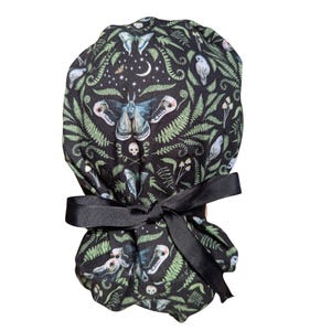 May include: A black scrub cap with a repeating pattern of moths, skulls, ferns, and celestial elements. The cap is tied with a black ribbon. The design includes shades of green, blue, and white.