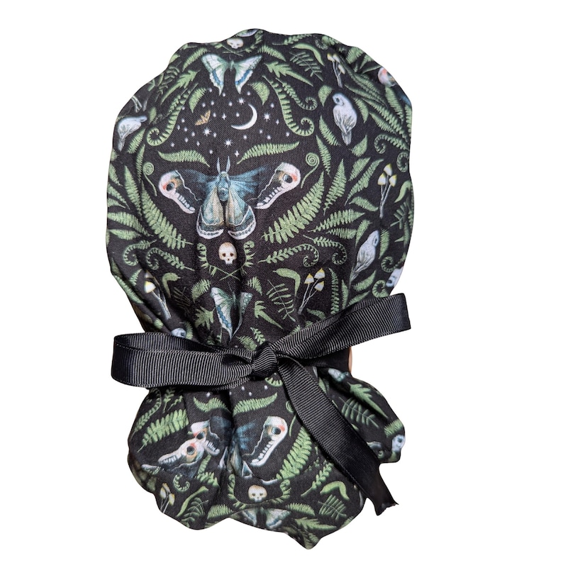 May include: A black scrub cap with a repeating pattern of moths, skulls, ferns, and celestial elements. The cap is tied with a black ribbon. The design includes shades of green, blue, and white.