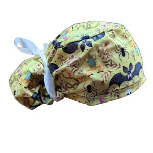 May include: A yellow scrub cap with a Halloween-themed pattern. The fabric features black bats, spiders, snakes, and a frog, along with floral and leaf accents. A white ribbon is attached for tying.