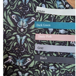 May include: Close-up of a fabric with a dark background featuring a moth and fern pattern. Ribbons in white, dark green, dusty pink, gray, and black are displayed, each labeled with its color.