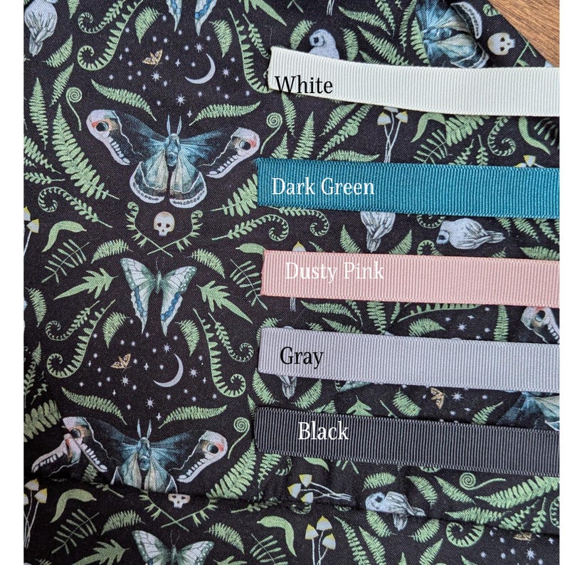 May include: Close-up of a fabric with a dark background featuring a moth and fern pattern. Ribbons in white, dark green, dusty pink, gray, and black are displayed, each labeled with its color.
