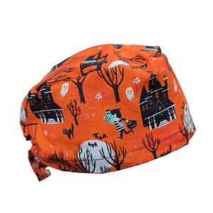 May include: Orange scrub hat with a Halloween-themed print featuring black houses, trees, ghosts, and a character dressed in a black and white striped shirt and carrying a bag of candy.