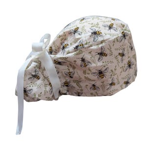 May include: A light pink scrub cap featuring a repeating pattern of bees and green foliage. The cap has a white ribbon tie closure. The bees are black and yellow, and the foliage is a muted green.