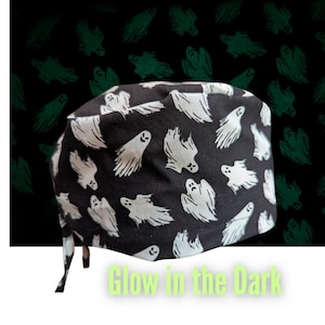 May include: Black and white patterned surgical cap featuring a repeating ghost design. The cap has ties for a secure fit. The words "Glow in the Dark" are in a light green color at the bottom of the image. The background has a repeating pattern of green ghosts.