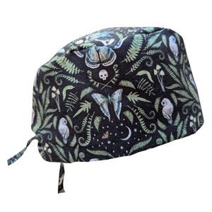 May include: A black surgical scrub cap with a whimsical pattern of butterflies, owls, mushrooms, ferns, and a skull. The cap has a tie closure and is made of a soft, patterned fabric. The design includes a crescent moon and stars.