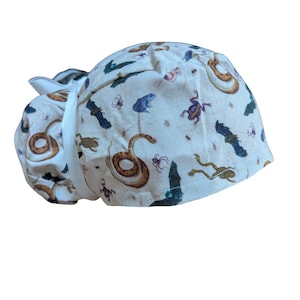 May include: A cream-colored surgical scrub cap featuring a repeating pattern of snakes, bats, frogs, and insects in shades of brown, blue, and purple. The cap has a white tie closure.